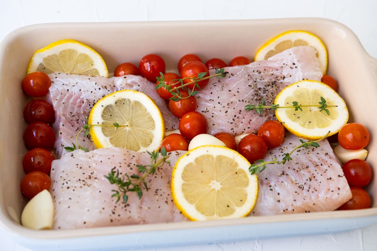 Catfish fillet, cherry tomatoes, and lemon slices arranged in a baking dish. 