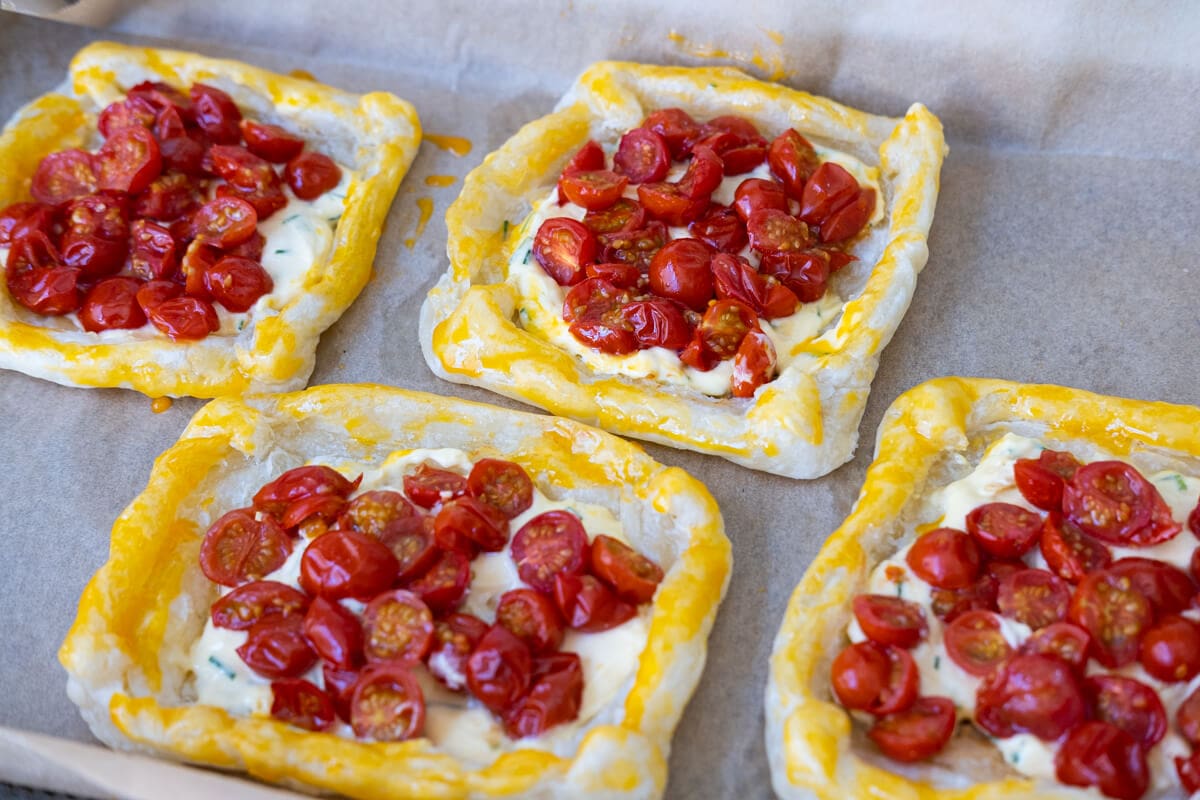 Four rectangle puff pastry sheet topped with roasted cherry tomato and with egg washed pastry borders  arranged on top of oven tray lined with baking paper.