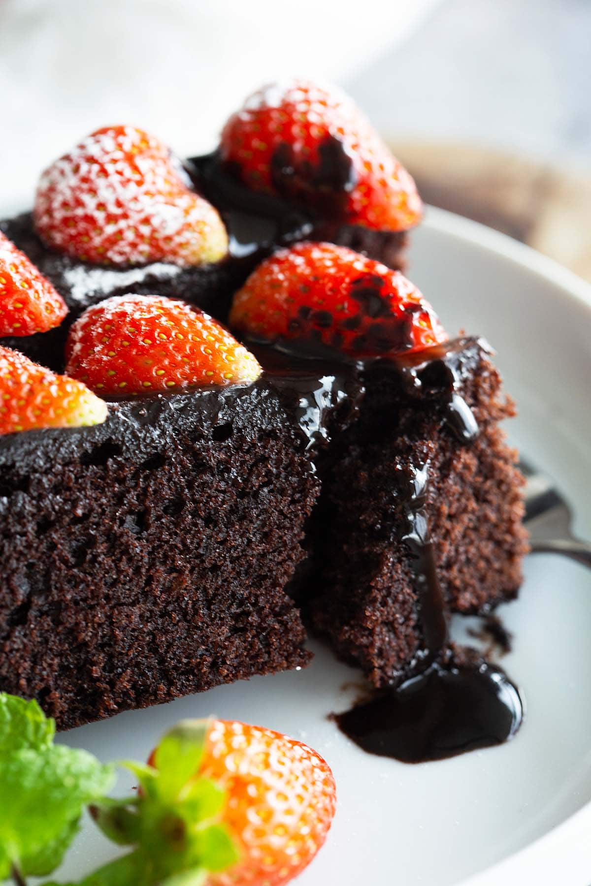 Pieces of moist chocolate cake with chocolate sauce dripping off and strawberries on top served in a plate. 