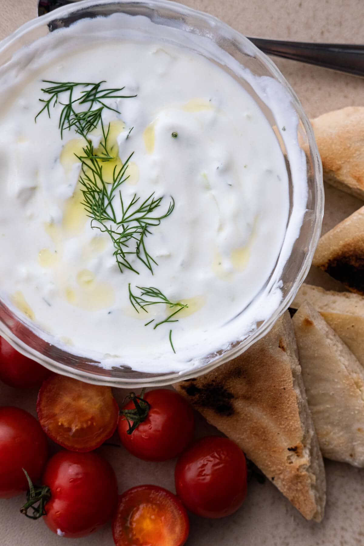 Creamy classic tzatziki sauce topped with fresh dill and olive oil drizzle served in a bowl with tomatoes and pita bread at the side. 