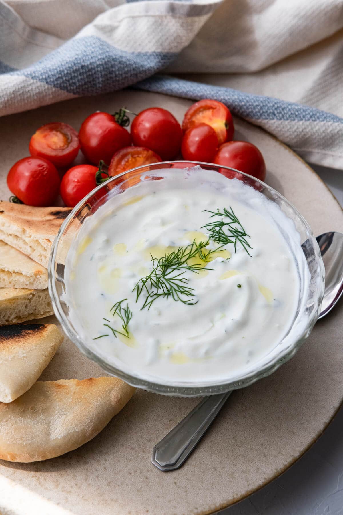White Tzatziki sauce topped with thyme served in a small bowl alongside cherry tomatoes and pita bread. 