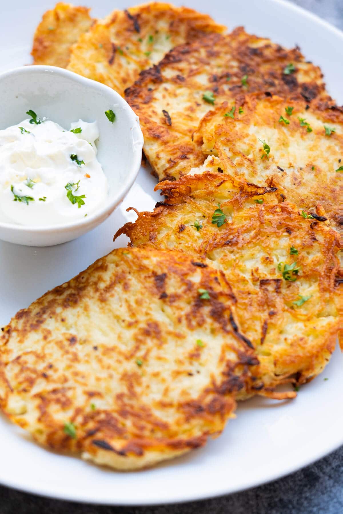 Golden brown potato pancakes lined around a bowl of sour cream on a white plate. 
